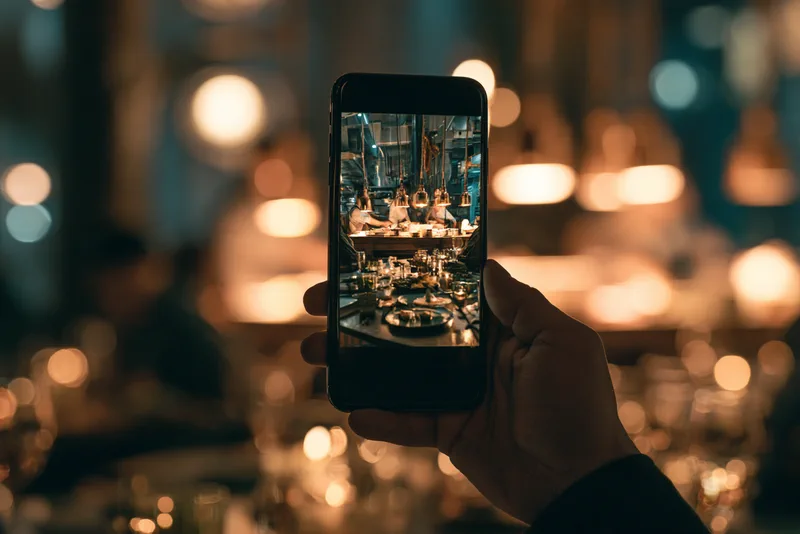 Service — a hand holding a phone up in a candlelit dining room, the screen showing the pass of the restaurant's kitchen