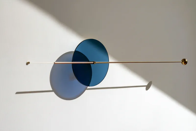 Tide Clock — a slim brass rod mounted on a white wall carrying two overlapping discs of blue glass, their shadows falling across the wall