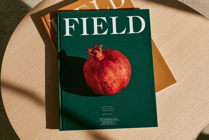 Field, Redesigned — two copies of Field magazine on a pale oak table, the top cover a single pomegranate on deep green under the masthead FIELD in white capitals
