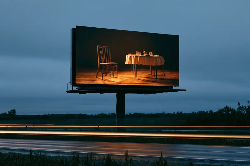The Empty Chair — a roadside billboard at dusk showing an empty chair pulled back from a laid table, traffic passing as streaks of light