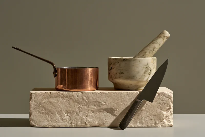 Ten Objects for a Kitchen — a still life of a copper saucepan, a marble mortar and pestle and a kitchen knife arranged on a block of rough stone