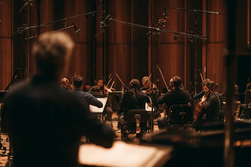 Score for Strings and Tape — a string orchestra mid-session in a wood-panelled recording studio, seen over the conductor's shoulder